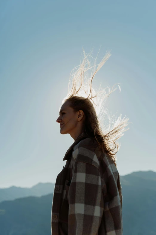 a woman with her hair blowing in the wind, by Matthias Weischer, trending on unsplash, clear sky, with mountains in background, back lit, sun behind him