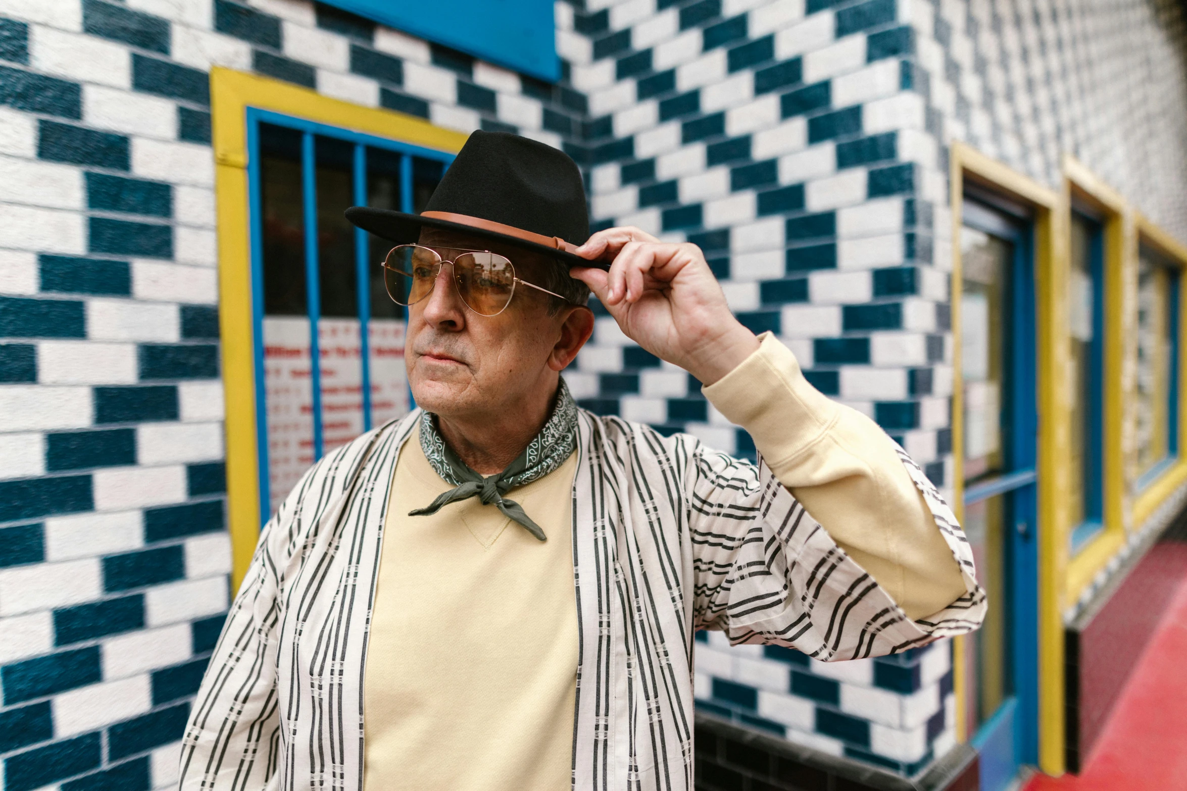 a man wearing a hat standing in front of a building, an album cover, inspired by Wyndham Lewis, unsplash, bauhaus, patterned clothing, an oldman, with yellow cloths, al pacino