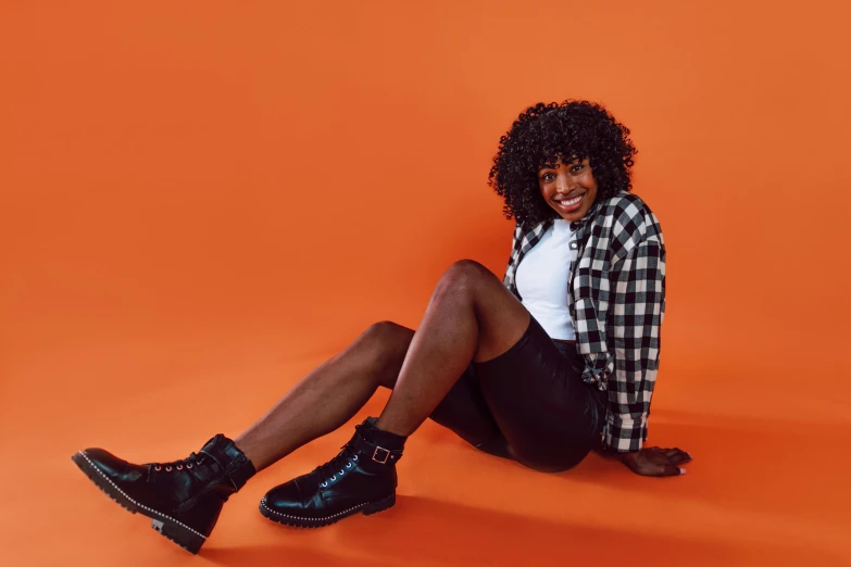 a woman sitting on the ground with her legs crossed, trending on pexels, in front of an orange background, super cute funky black girl, black leather boots, dark short curly hair smiling