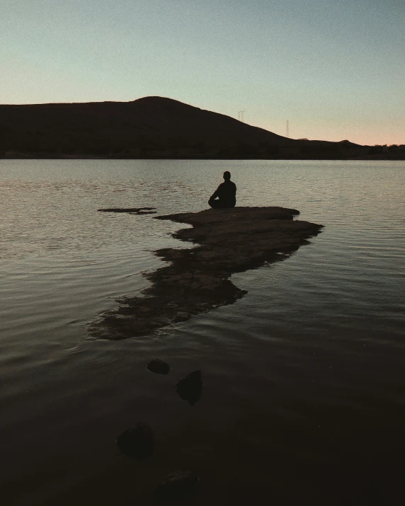 a person sitting on a rock in the middle of a lake, in the evening