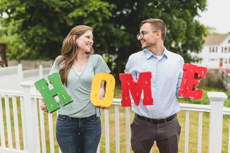 a man and woman standing next to each other holding the word home, a colorized photo, unsplash, letterism, doug walker, rainbow accents, profile image, fan favorite