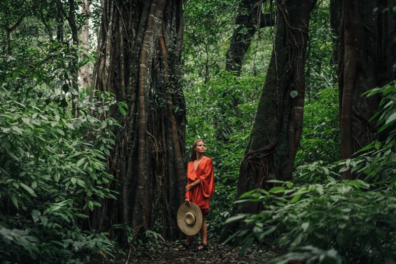 a monk standing in the middle of a forest, by Caro Niederer, sumatraism, wearing an orange jumpsuit, red dress and hat, conde nast traveler photo, lush greenery