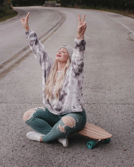a woman sitting on a skateboard in the middle of the road, by Kristin Nelson, trending on pexels, pose(arms up + happy), grey sweater, tie-dye, peace sign