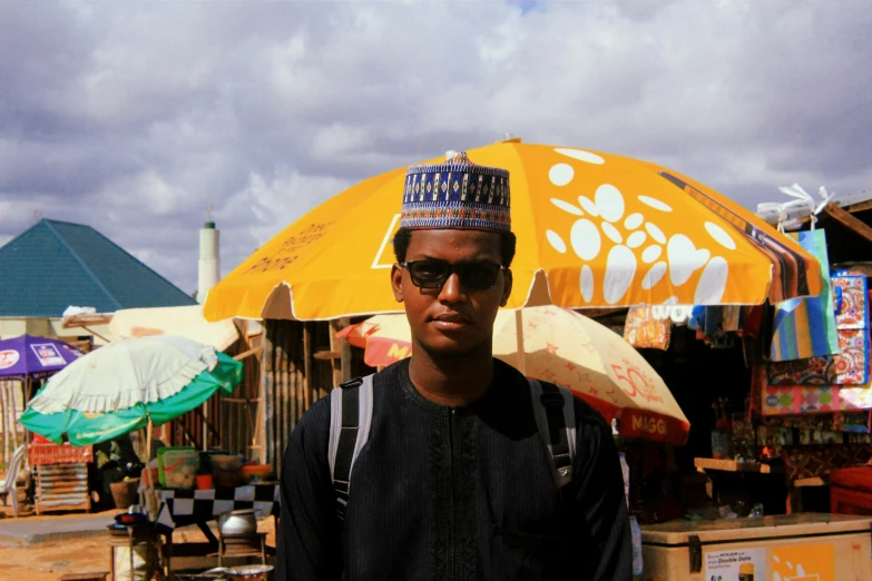 a man standing in front of a yellow umbrella, hurufiyya, wearing sunglasses and a hat, somali attire, light-brown skin, market setting