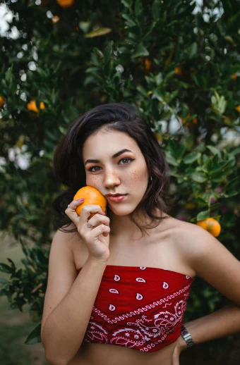 a woman holding an orange in front of a tree, by Robbie Trevino, pexels, 2 4 year old female model, half asian, 🐿🍸🍋, 5 0 0 px models