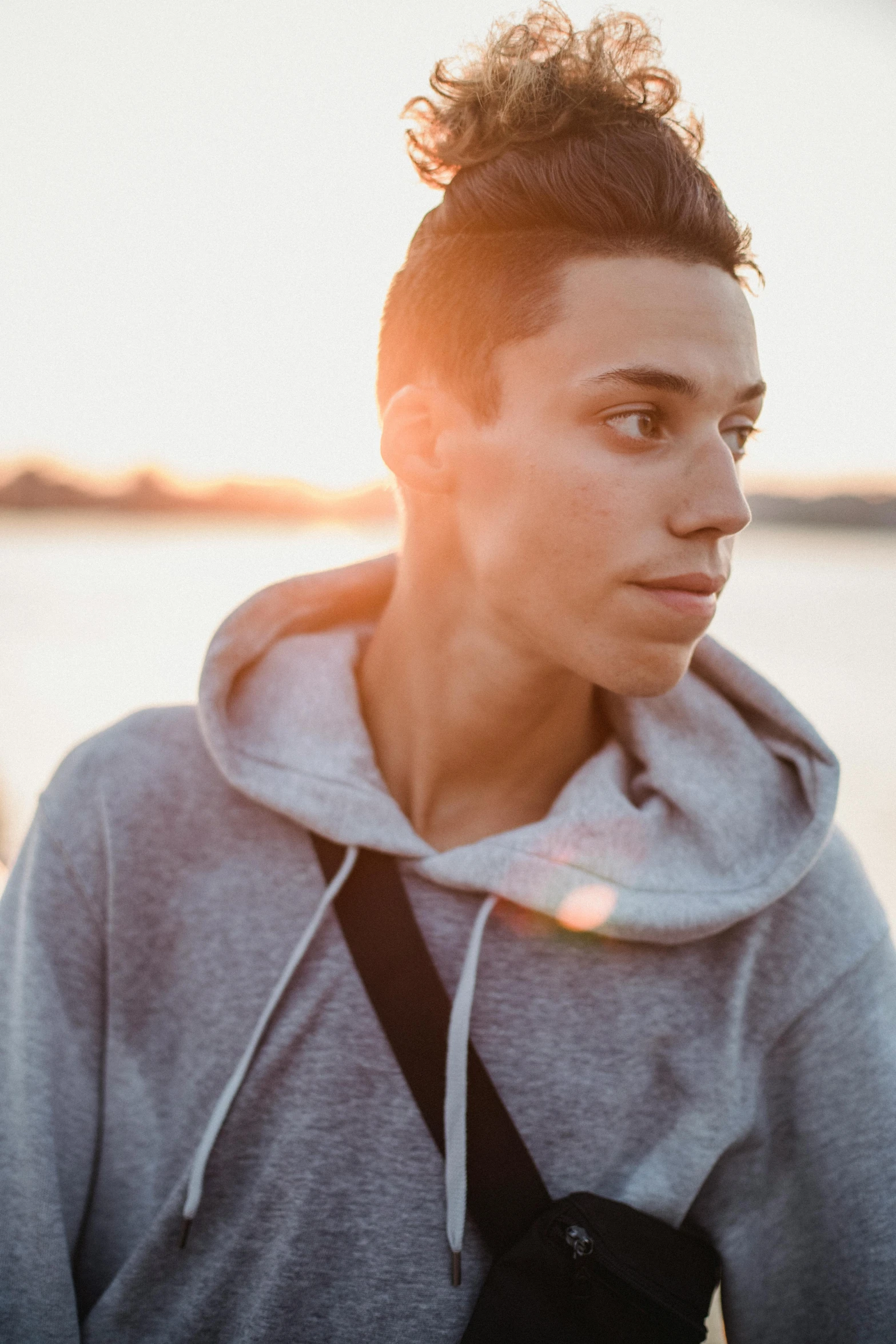 a woman standing in front of a body of water, by Jacob Toorenvliet, trending on unsplash, antipodeans, portrait of beautiful young man, wearing a hoody, pete davidson, as the sun sets on the horizon