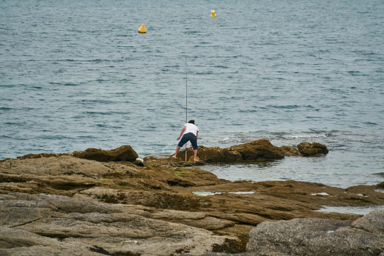 a man standing on top of a rock next to the ocean, fishing pole, manly, rocky seashore, wheres wally