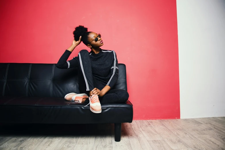 a woman sitting on a couch in front of a red wall, by Lily Delissa Joseph, trending on pexels, black teenage girl, wearing fitness gear, leather couch, pink and black