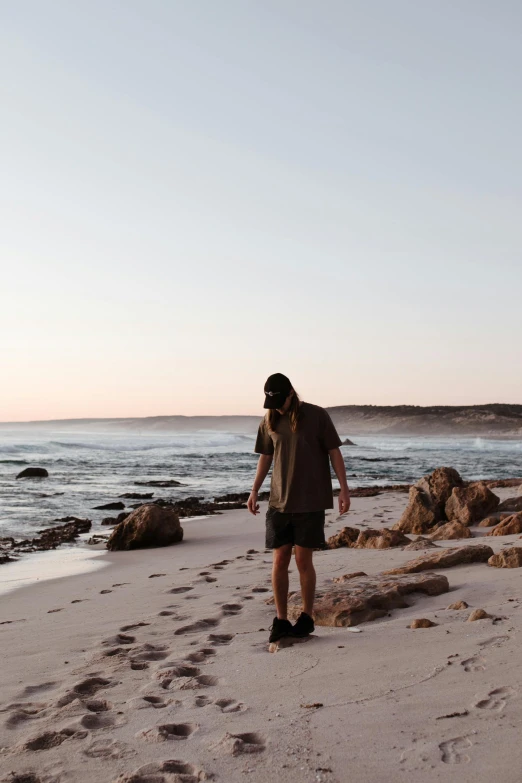 a man standing on top of a sandy beach next to the ocean, by Lee Loughridge, trending on pexels, aboriginal australian hipster, summer evening, standing on rocky ground, walking away