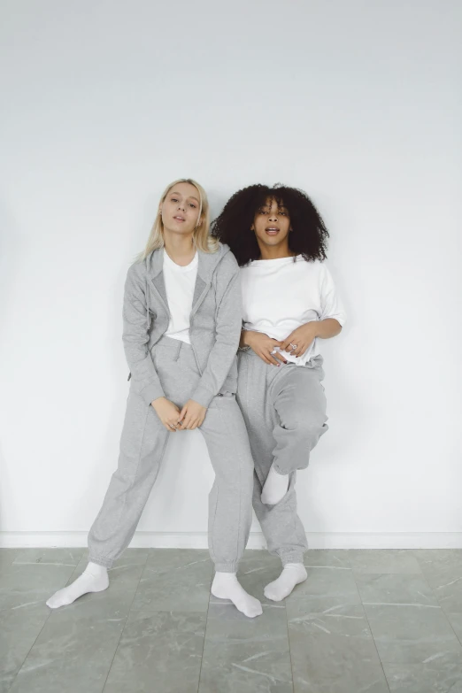 two women standing next to each other in front of a white wall, trending on pexels, wearing a hoodie and sweatpants, made in tones of white and grey, promotional image, roomies