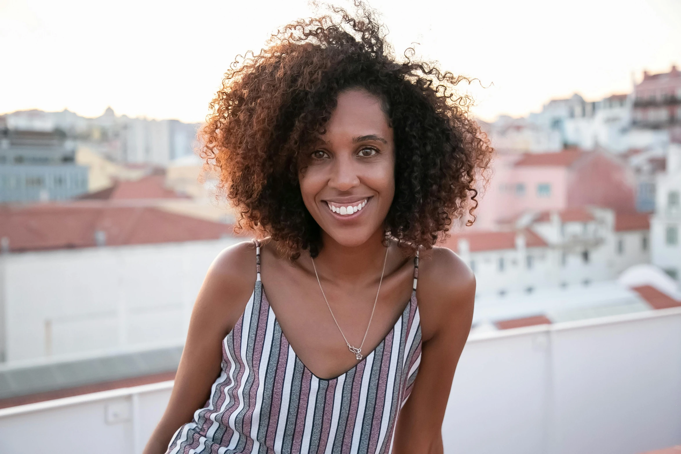 a beautiful young woman sitting on top of a roof, by Almada Negreiros, pexels contest winner, happening, dark short curly hair smiling, ( ( brown skin ) ), closeup headshot, portugal