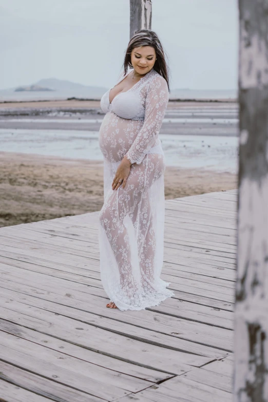 a pregnant woman standing on a boardwalk at the beach, pexels contest winner, happening, wearing translucent sheet, intricate details. front on, robe, maternal