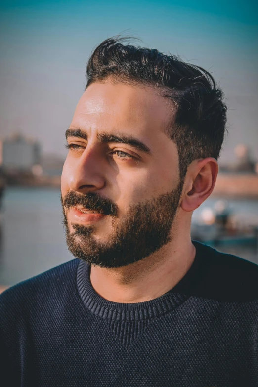 a man standing in front of a body of water, a picture, inspired by Ahmed Yacoubi, headshot profile picture, discord profile picture, shaved beard, profile image