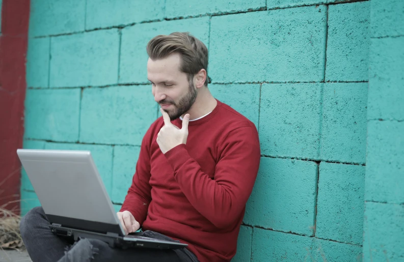 a man sitting against a wall using a laptop, pexels, teal silver red, curious, bearded, wearing red