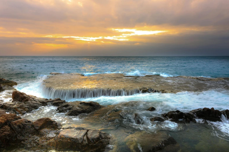 a large body of water sitting on top of a rocky beach, by Elizabeth Durack, pexels contest winner, water cascading, manly, golden rays, sea foam