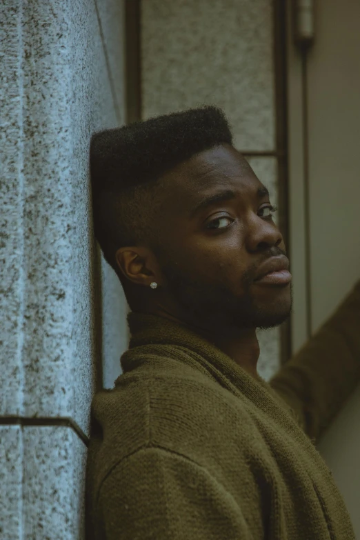 a man in a bathrobe leaning against a wall, an album cover, inspired by Theo Constanté, pexels contest winner, realism, jaylen brown, lgbtq, he is wearing a brown sweater, pompadour