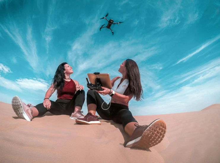 a couple of women sitting on top of a sandy beach, a picture, pexels contest winner, realism, hovering drone, avatar image, electronics, riding in the sky