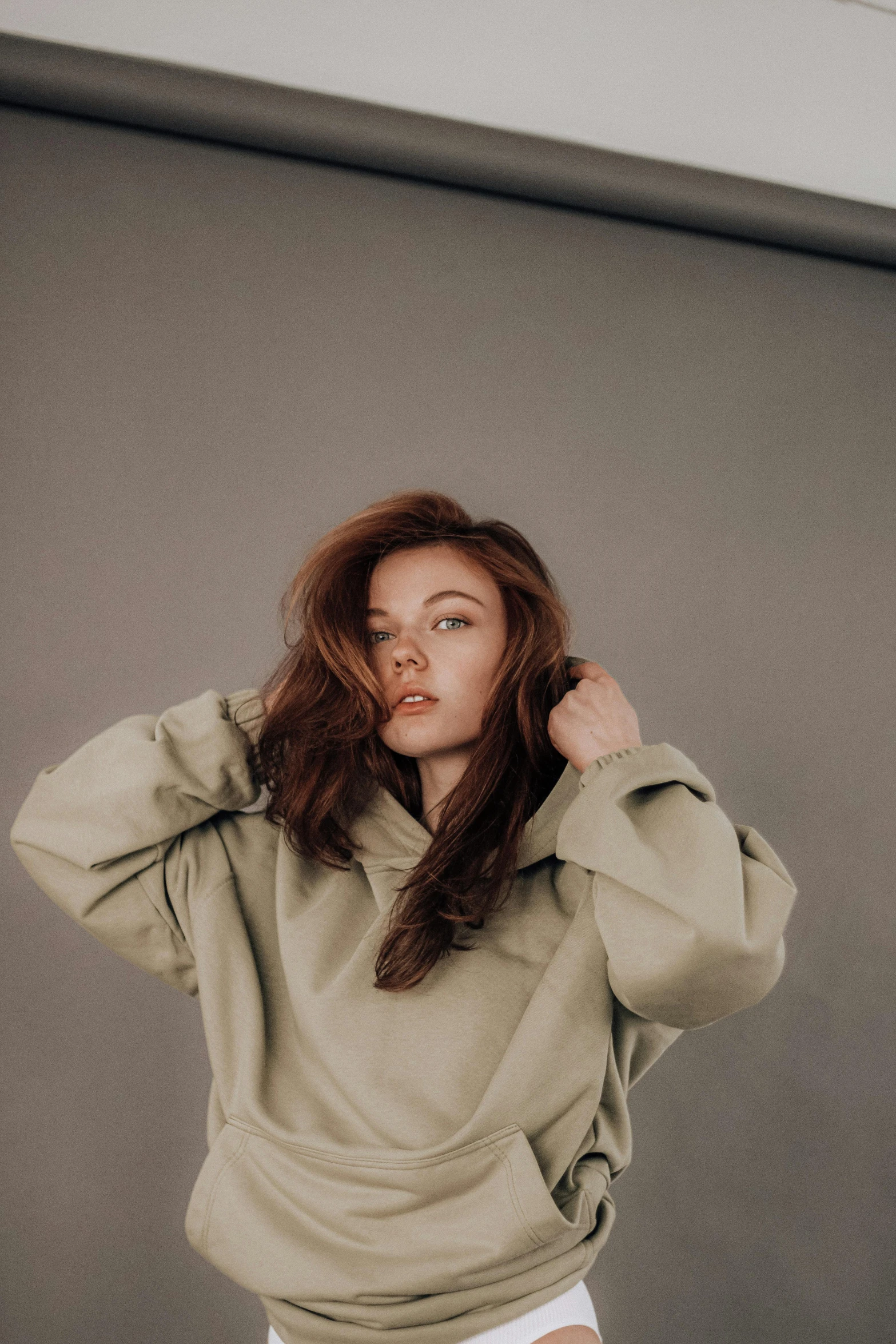 a woman standing in front of a gray wall, inspired by Elsa Bleda, trending on pexels, renaissance, beige hoodie, ( redhead, charli bowater, gif
