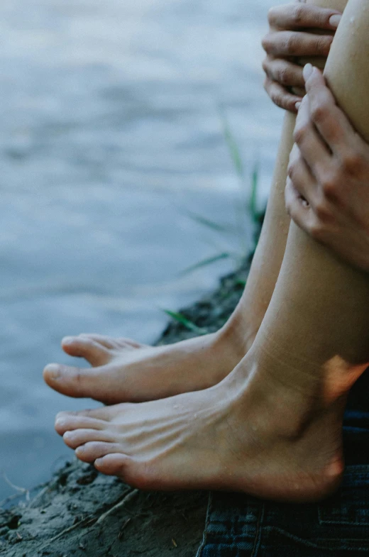 a person sitting on a rock next to a body of water, closeup of arms, barefeet, flirting, dry skin