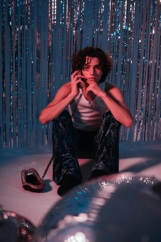 a woman sitting on the ground talking on a cell phone, an album cover, joe keery, neon lights in the background, curls on top of his head, standing in a dimly lit room