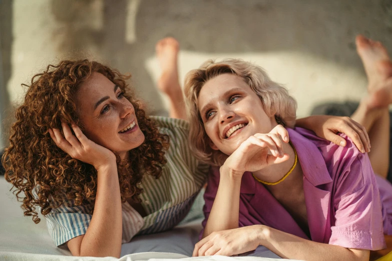 a couple of women laying on top of a bed, trending on pexels, short curly blonde haired girl, smiling at each other, retro stylised, good friends
