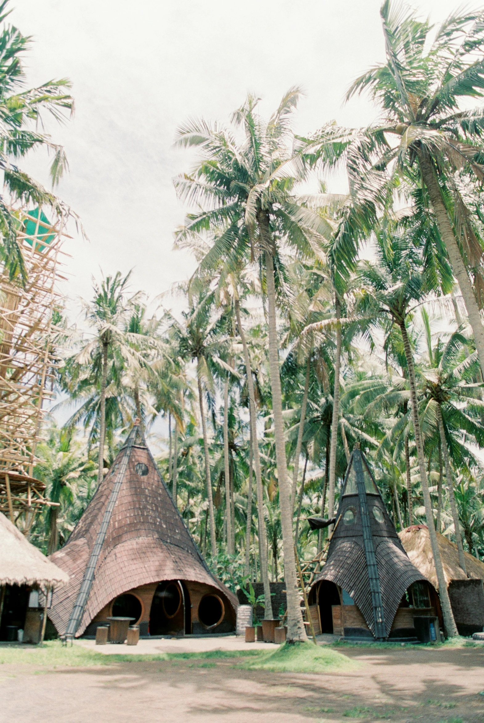 a group of huts sitting on top of a lush green field, inspired by Károly Kisfaludy, sumatraism, palm trees on the beach, tall stone spires, festival vibes, exterior