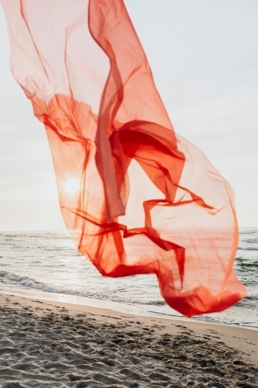 a woman standing on top of a sandy beach next to the ocean, inspired by Christo, unsplash contest winner, conceptual art, swirling red-colored silk fabric, diaphanous translucent cloth, detail shot, on a gray background