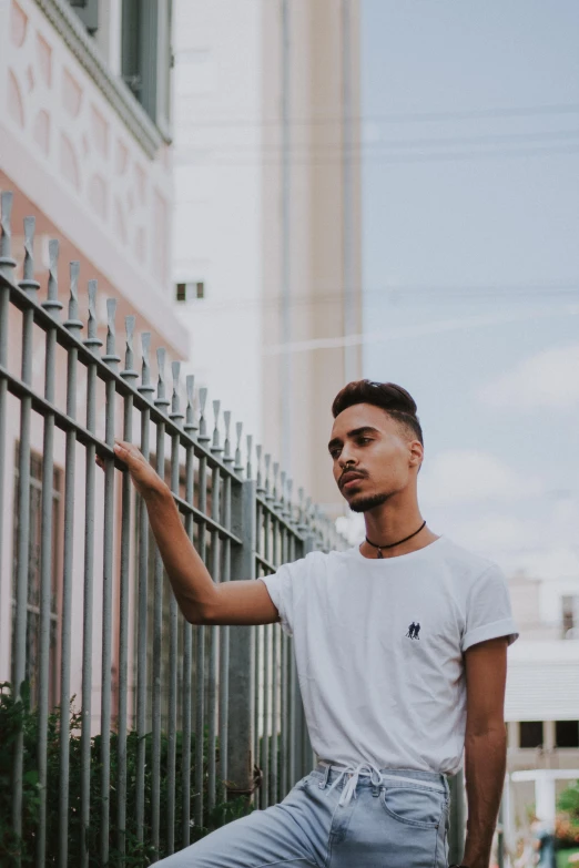 a man leaning against a fence in front of a building, by Robbie Trevino, androgynous person, dressed in a white t shirt, smooth background, khyzyl saleem