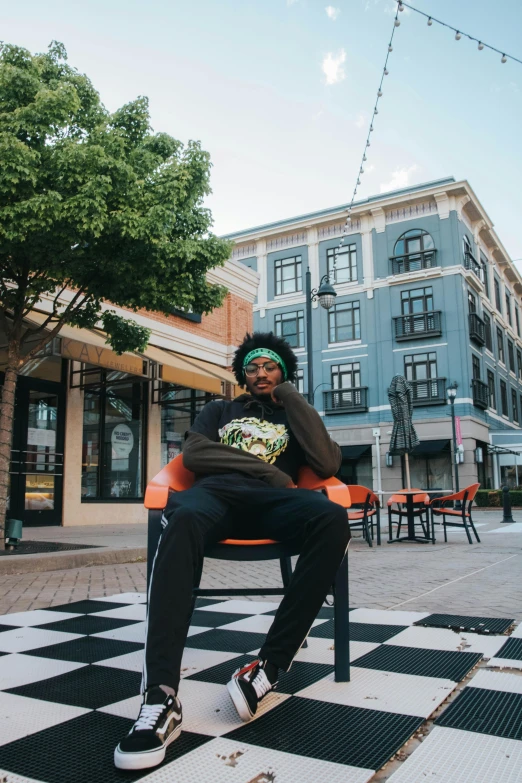 a person sitting on a bench in front of a building, teddy fresh, african canadian, in town, vibrant setting