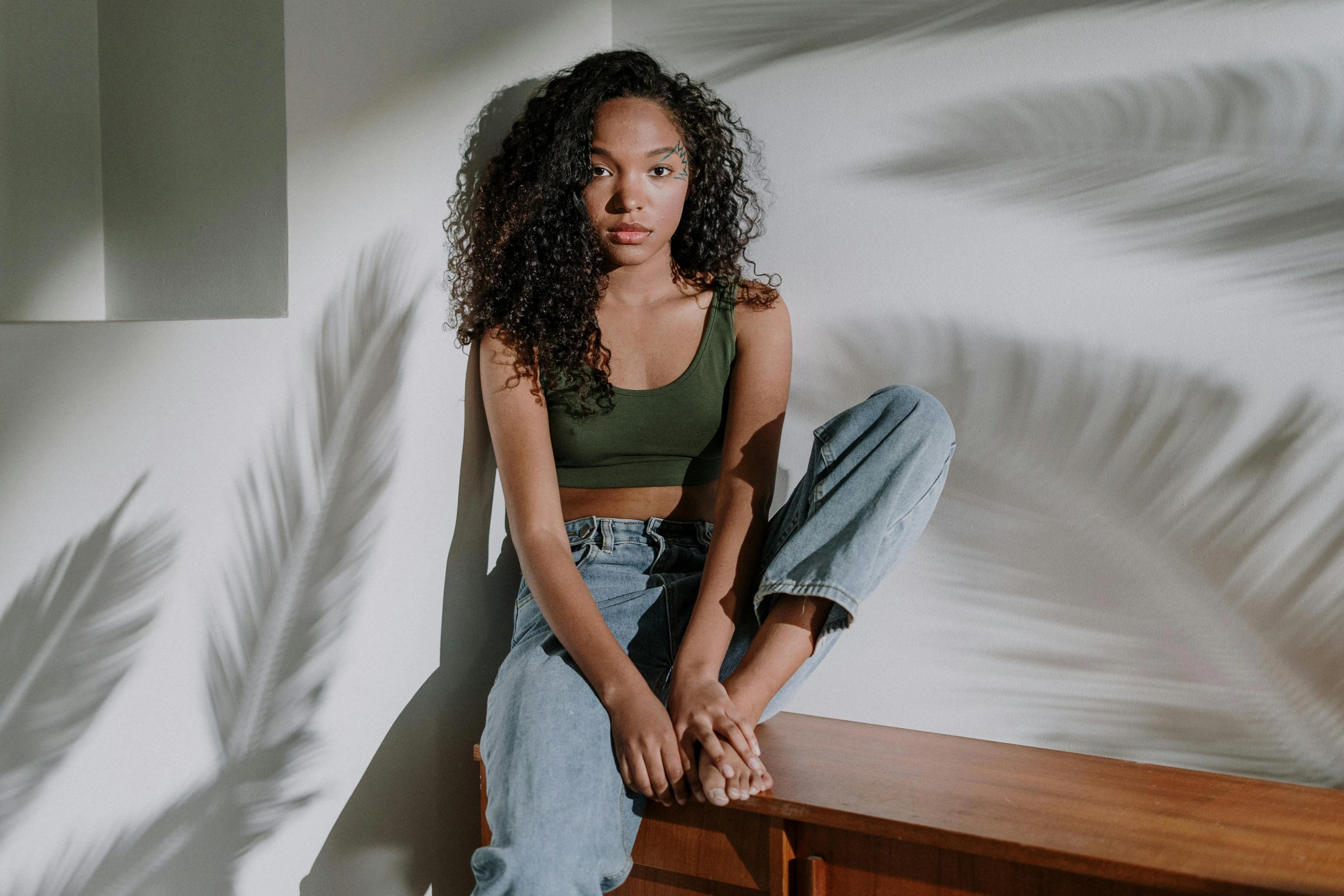 a woman sitting on top of a wooden bench, by Stokely Webster, trending on pexels, renaissance, green halter top, teenage vanessa morgan, leaning against the wall, curly hair