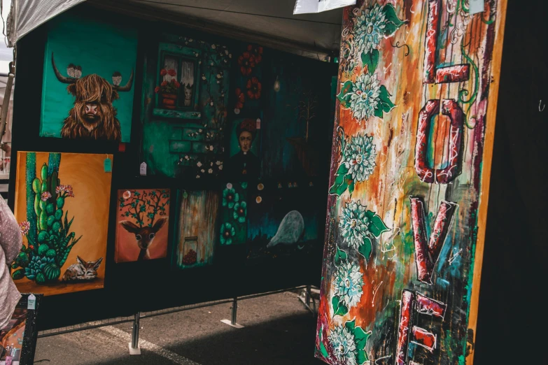 a woman standing in front of a booth with paintings on it, pexels contest winner, street art, love art, street of teal stone, thumbnail, market stalls