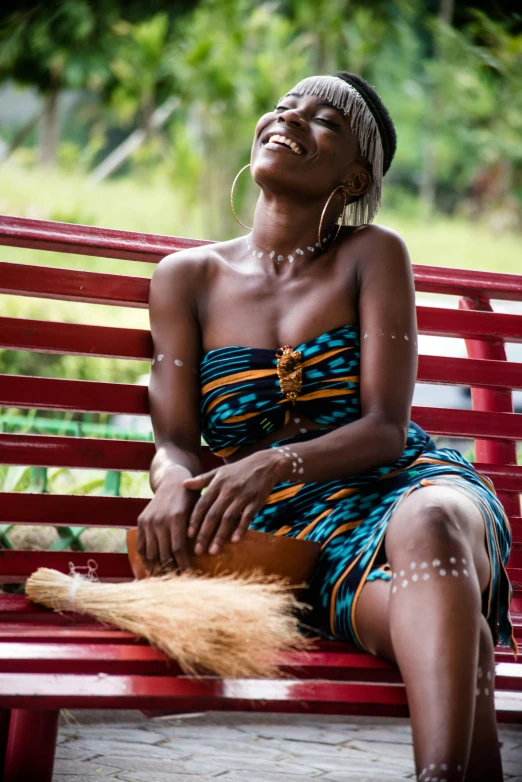 a woman sitting on top of a red bench, by Ingrida Kadaka, pexels contest winner, afrofuturism, facial tribal markings, tan skin, smiling playfully, wearing a dress made of beads