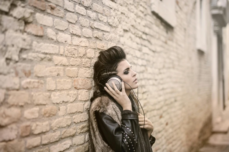 a woman leaning against a brick wall talking on a cell phone, an album cover, inspired by Elsa Bleda, trending on pexels, giant headphones rocking out, wearing studded leather, brunette, alessio albi