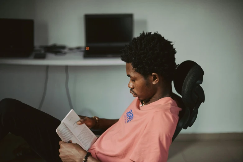 a woman sitting in a chair reading a book, by Lily Delissa Joseph, pexels contest winner, afrofuturism, black teenage boy, in the office, a young man, in a chill position