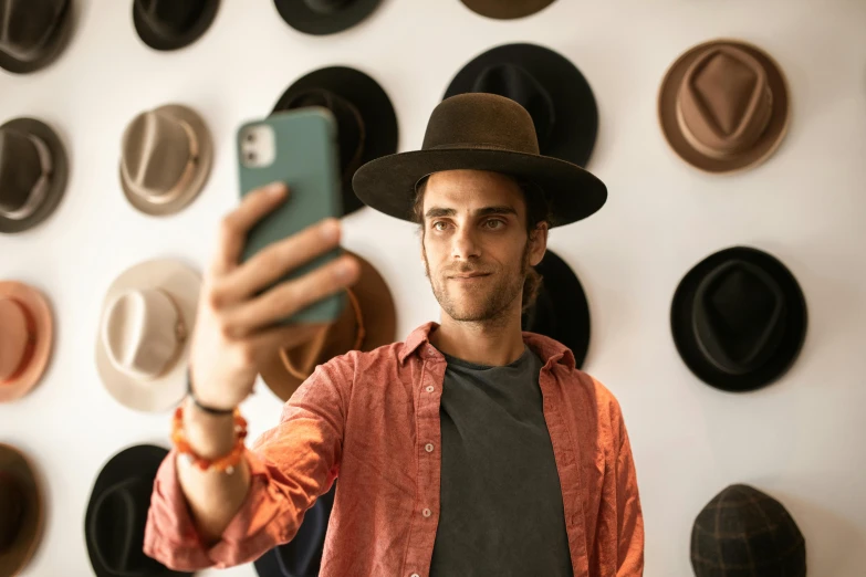 a man taking a selfie in front of a wall of hats, trending on pexels, young spanish man, avatar image, australian, caracter with brown hat