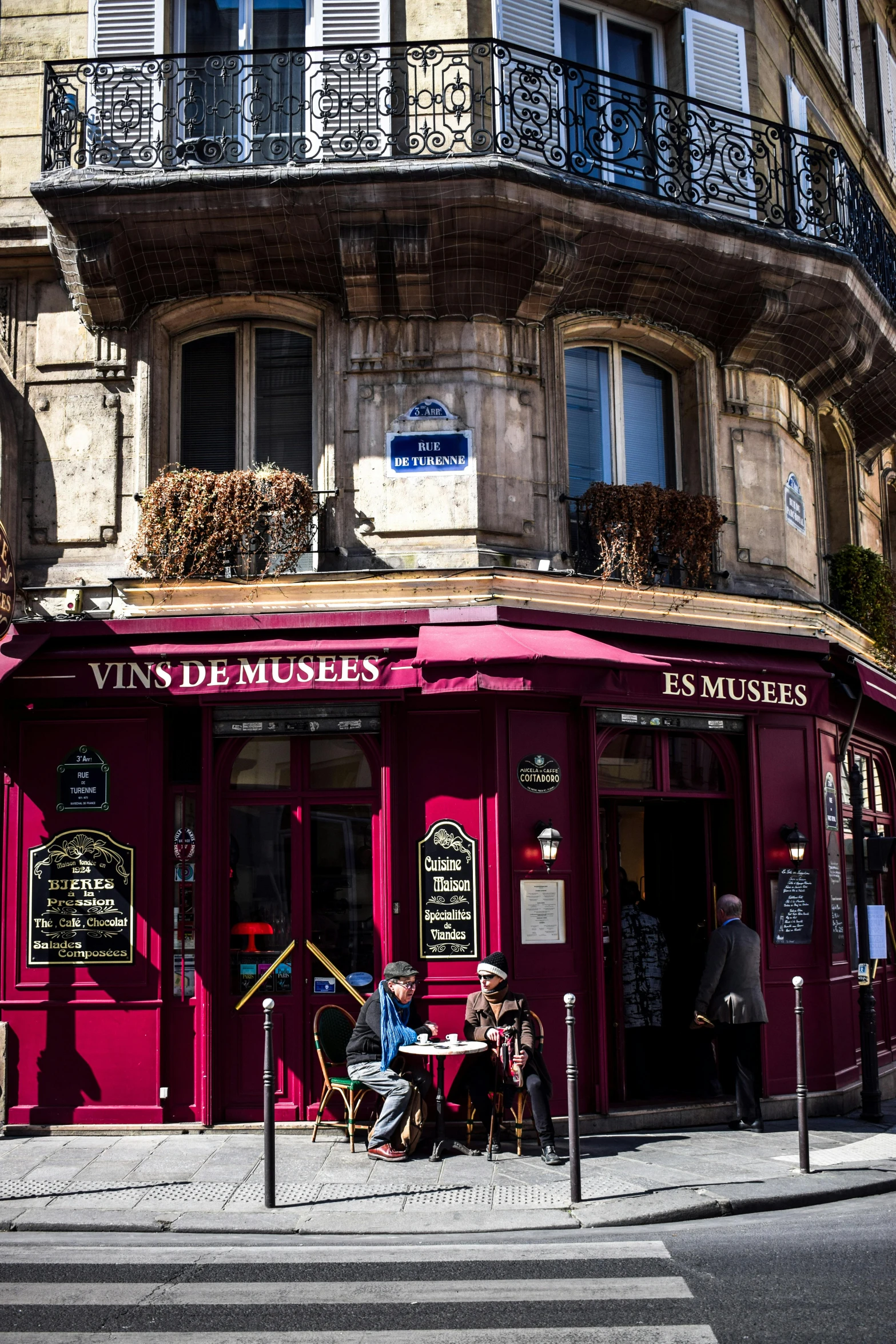 a couple of people that are sitting outside of a building, art nouveau, wine-red and grey trim, very smoky paris bar, colorful signs, museum