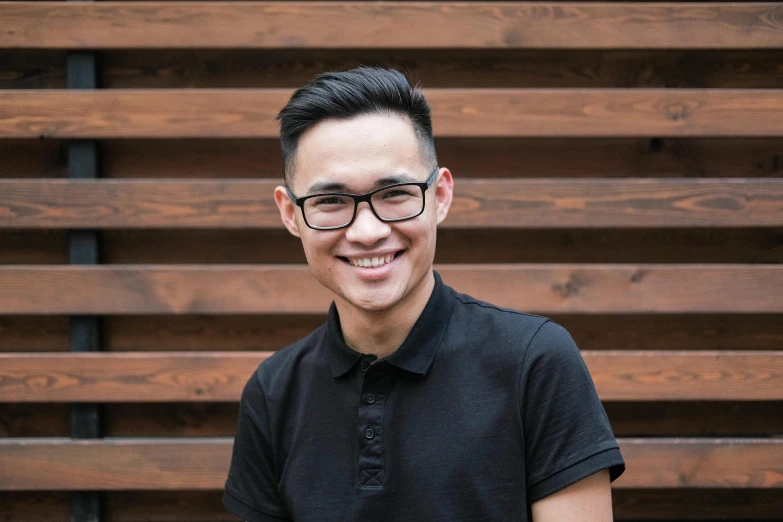 a man standing in front of a wooden wall, inspired by Patrick Ching, wearing black rimmed glasses, darren quach, friendly smile, avatar image