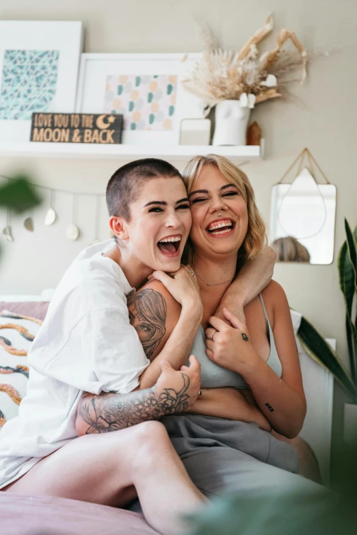 a woman sitting on top of a bed next to a man, a picture, trending on pexels, antipodeans, brown buzzcut, lesbian embrace, both laughing, with tattoos