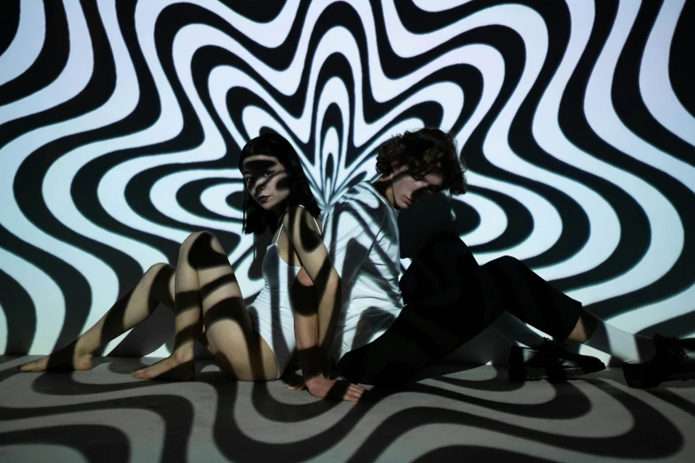 a couple of people sitting on top of a floor, video art, infinite psychedelic waves, white stripes all over its body, light and dark, posed