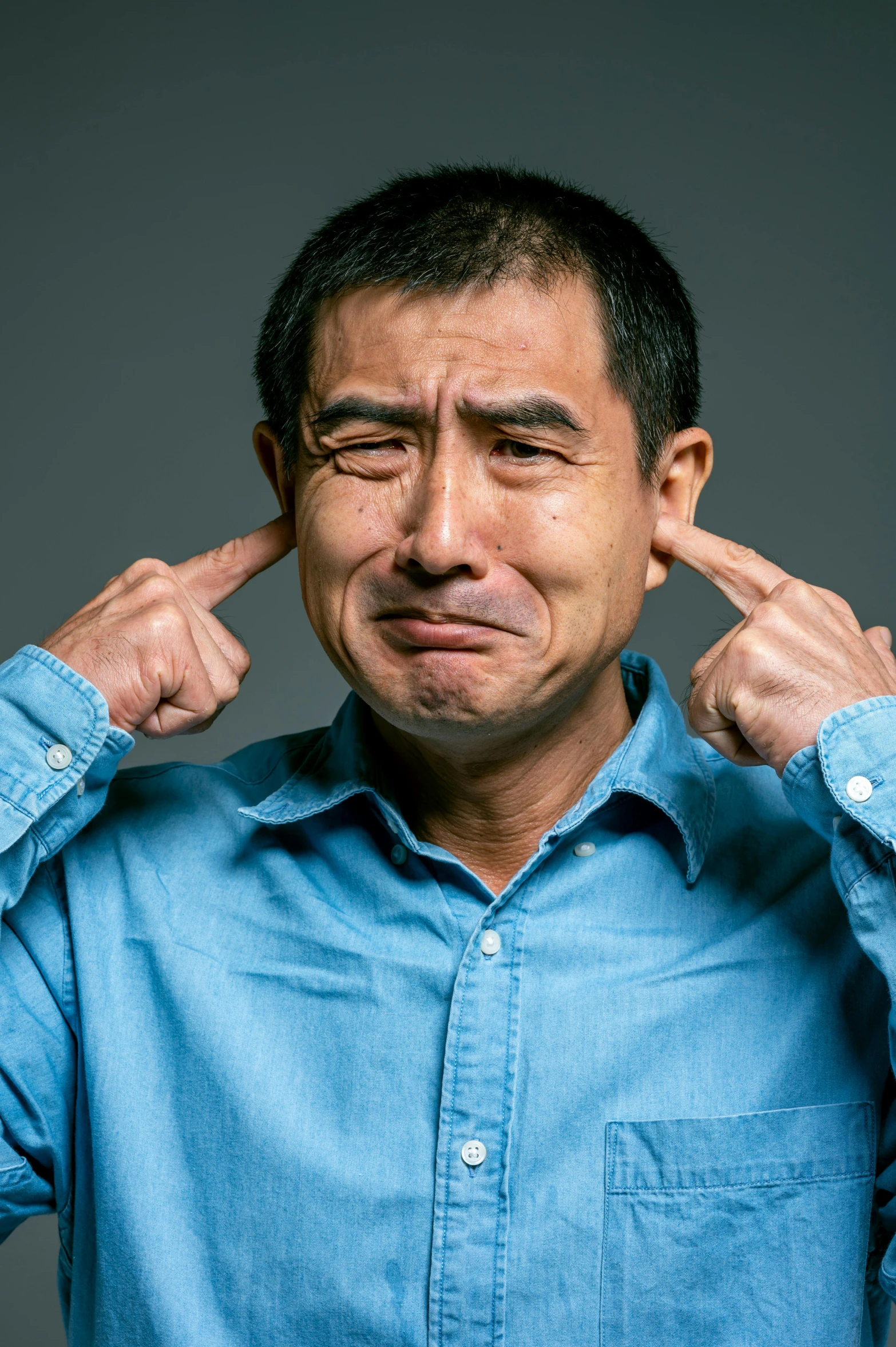 a man in a blue shirt holding his hands to his ears, by Miyamoto, shutterstock, mingei, frowning expression, instagram post, ethnicity : japanese, naoto hattori