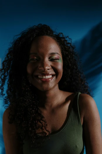 a woman with curly hair smiling at the camera, teal studio backdrop, dark skinned, with the sun shining on it, pose 4 of 1 6