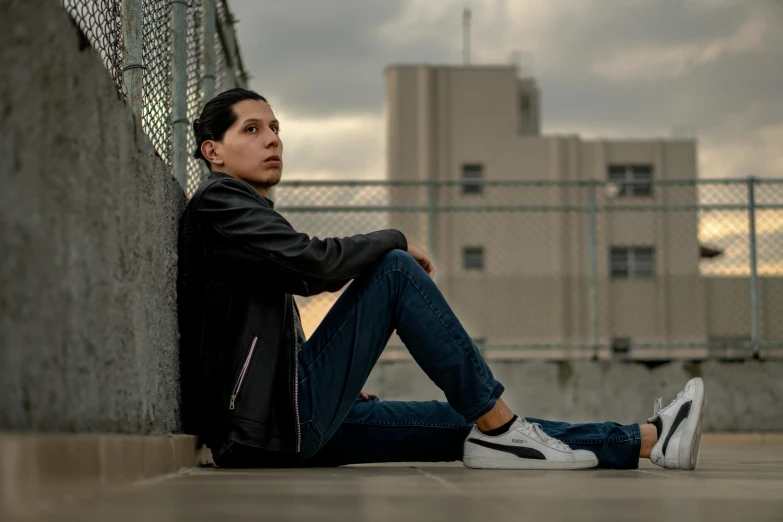 a man sitting on the ground in front of a fence, a portrait, inspired by Carlos Berlanga, pexels, androgynous male, standing on rooftop, high soles, concerned