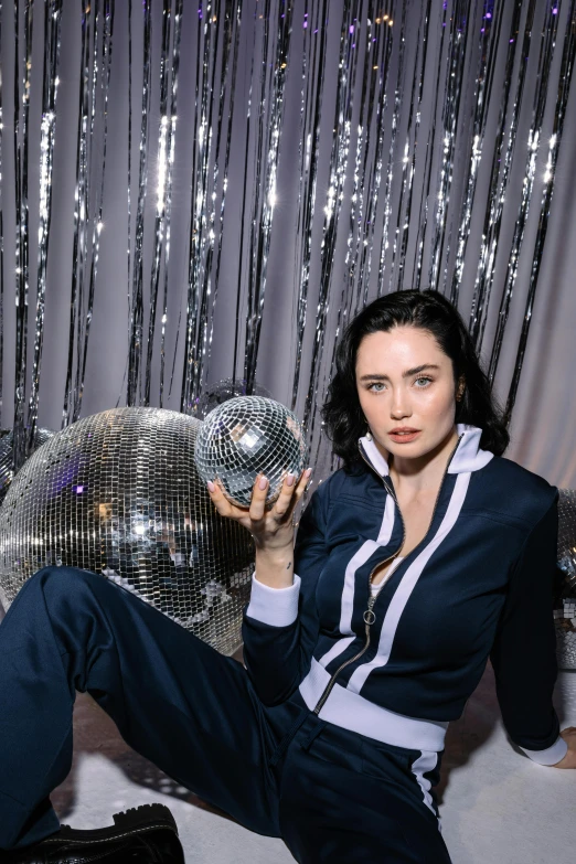 a woman sitting on a bed holding a disco ball, woman with black hair, wearing a track suit, meredit frampton style, promo image