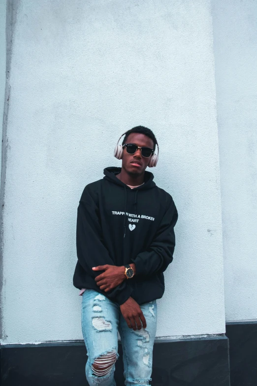 a man in a black hoodie leaning against a white wall, an album cover, inspired by Barthélemy Menn, trending on pexels, wearing shades, profile image, androgynous person, dark skin tone