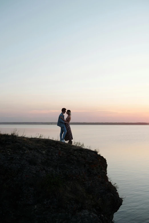 a couple standing on top of a cliff next to a body of water, a picture, unsplash, romanticism, rostov, late summer evening, kissing, russia