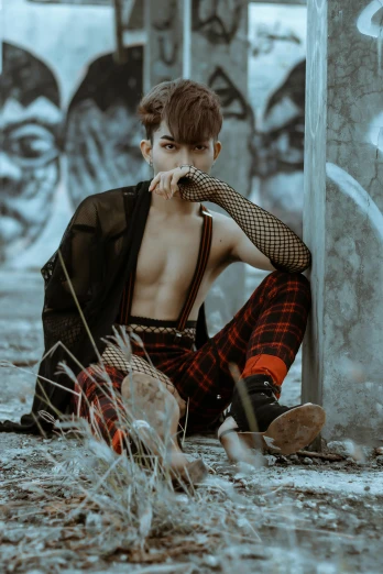 a man sitting on the ground in front of a graffiti covered wall, an album cover, inspired by Kim Deuk-sin, trending on pexels, wearing loincloth, checkered spiked hair, pose model, androgynous vampire