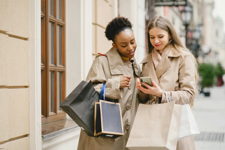 two women with shopping bags looking at a cell phone, by Matija Jama, trending on pexels, light brown trenchcoat, sustainable materials, thumbnail, amazing beauty