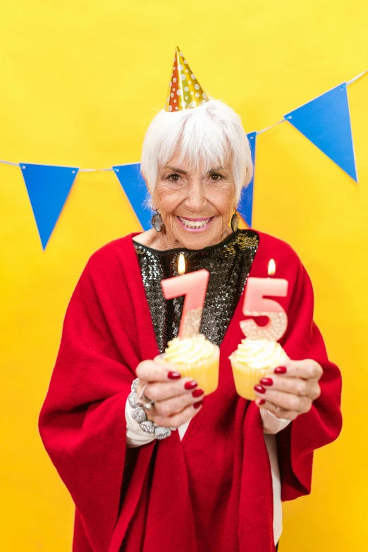 a woman standing in front of a yellow wall holding two cupcakes, a photo, by Juan O'Gorman, pexels, silver haired, happy birthday candles, wearing a king's cape, 7 5 mm