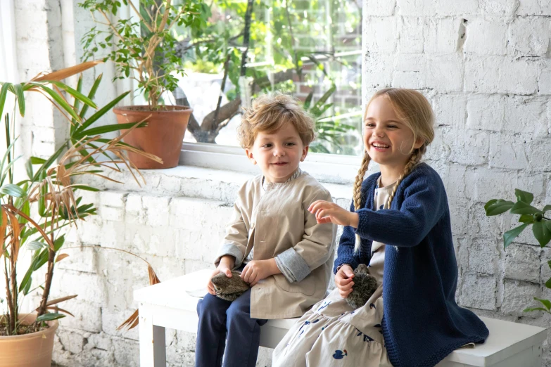 two children sitting on a bench in front of a window, a portrait, inspired by Elsa Beskow, unsplash, foliage clothing, navy, natural materials, smiling playfully