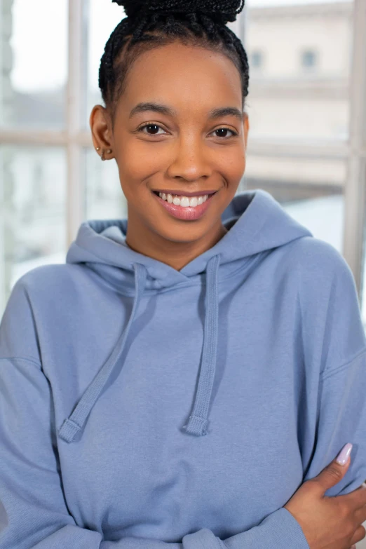 a woman in a blue hoodie standing in front of a window, inspired by Jean-Étienne Liotard, pexels contest winner, black teenage girl, happily smiling at the camera, promo image, loosely cropped